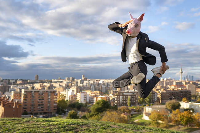 Empresário usando máscara de porco pulando na colina na cidade contra o céu — Fotografia de Stock