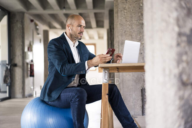 Бизнесмен с помощью мобильного телефона и ноутбука, сидя на фитнес-мяч в офисе — стоковое фото