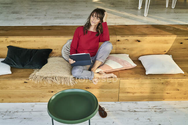 Женщина с рукой в волосах с помощью цифрового планшета, сидя в современном офисе — стоковое фото