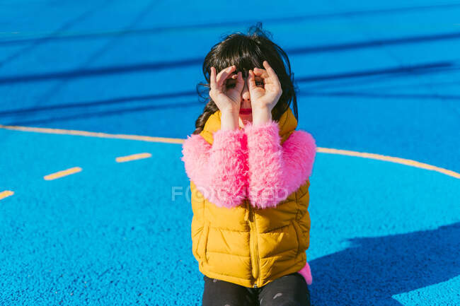 Chica formando prismáticos con los dedos mientras se sienta en la cancha deportiva - foto de stock