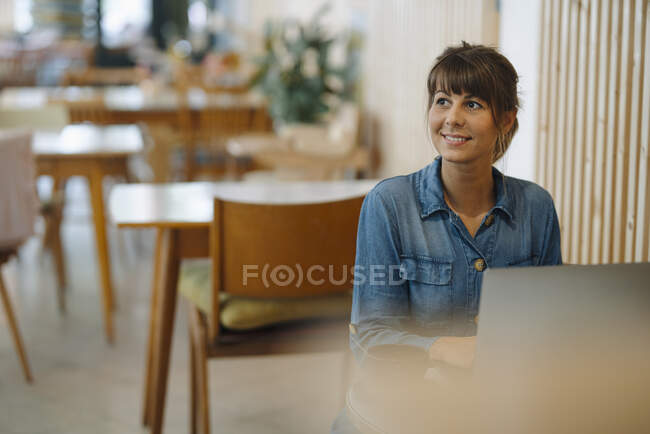 Бізнес-леді посміхається, дивлячись вбік, використовуючи ноутбук, сидячи в кав'ярні — стокове фото