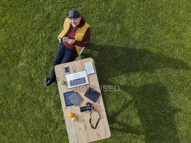 Aerial view of man sitting at coffee table set on green lawn and smiling up at camera with mug of coffee in hand — Stock Photo