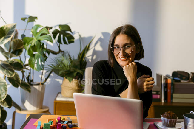 Empresária sorridente com a mão no queixo usando laptop enquanto está sentado em casa — Fotografia de Stock