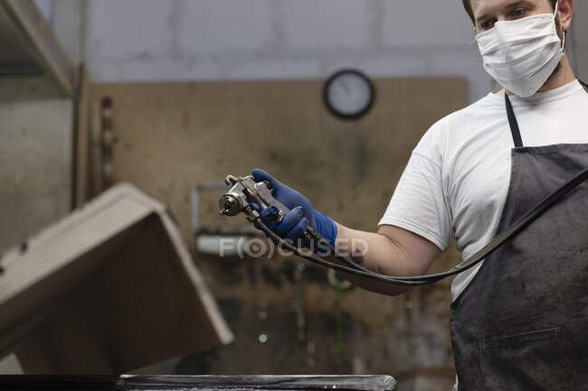 Man wearing face mask using spray paint gun on wood while standing at factory — Stock Photo