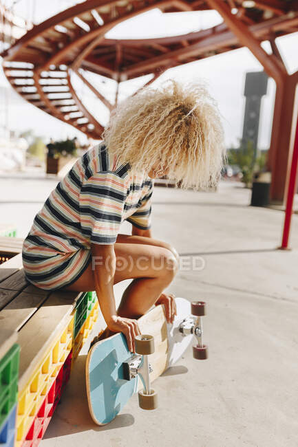 Середня доросла жінка з світлим кучерявим волоссям тримає скейтборд, сидячи на дерев'яному сидінні — стокове фото