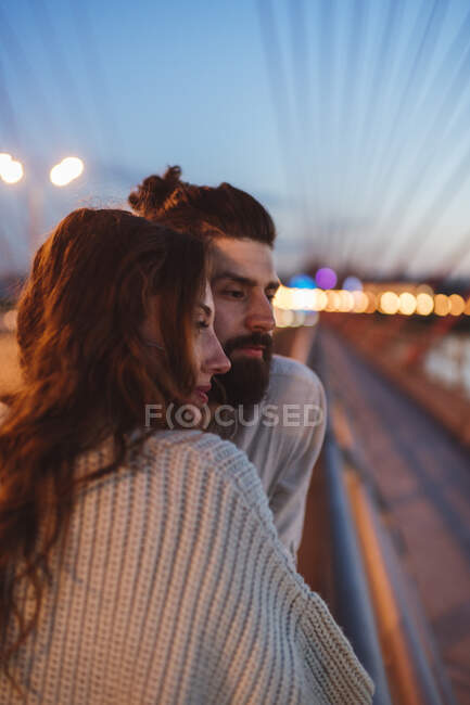 Продумана пара, дивлячись геть, стоячи на мосту в місті в сутінках — стокове фото