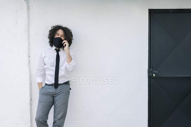 Homme d'affaires avec masque facial parlant sur téléphone portable tout en se tenant contre le mur — Photo de stock