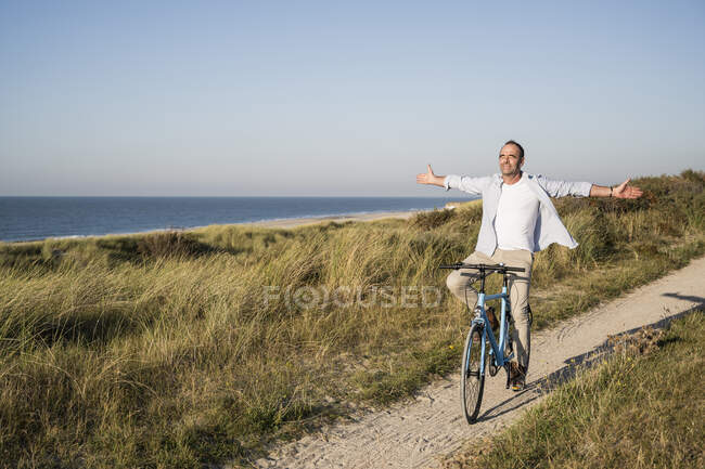 Homme mûr insouciant avec les bras tendus les yeux fermés tout en faisant du vélo à la plage — Photo de stock