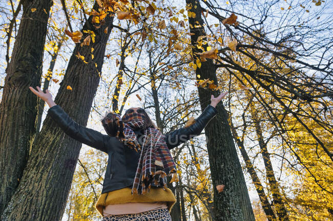 Mujer lanzando hojas secas de otoño mientras está de pie con los brazos extendidos contra los árboles en el parque - foto de stock