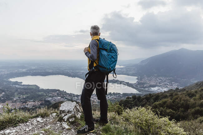 Зрелый мужчина с рюкзаком, смотрящий на озеро и город против неба на закате — стоковое фото
