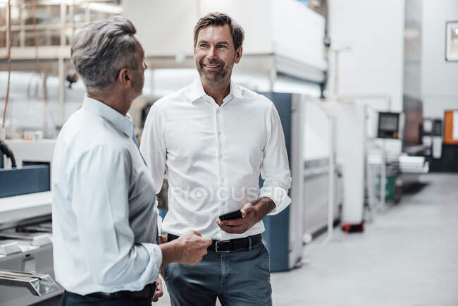Mature businessman holding smart phone while discussing with colleague in industry — Stock Photo