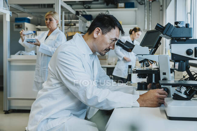 Scientifique homme assis au microscope tout en travaillant en arrière-plan au laboratoire — Photo de stock