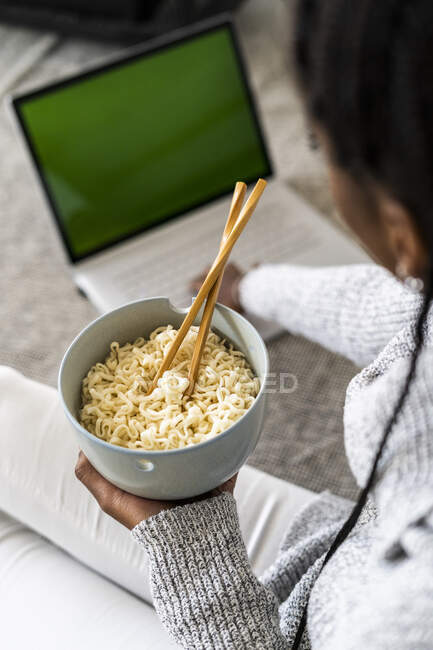 Жінка тримає миску з локшиною, використовуючи ноутбук вдома — стокове фото