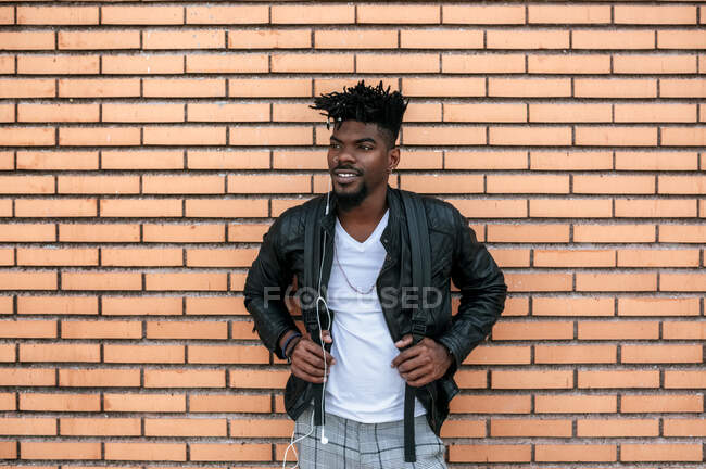 Hombre africano con chaqueta de cuero escuchando música mientras está de pie contra la pared de ladrillo - foto de stock