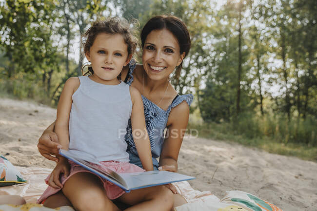 Lächelnde Mutter und Tochter lesen Buch, während sie am Strand sitzen — Stockfoto