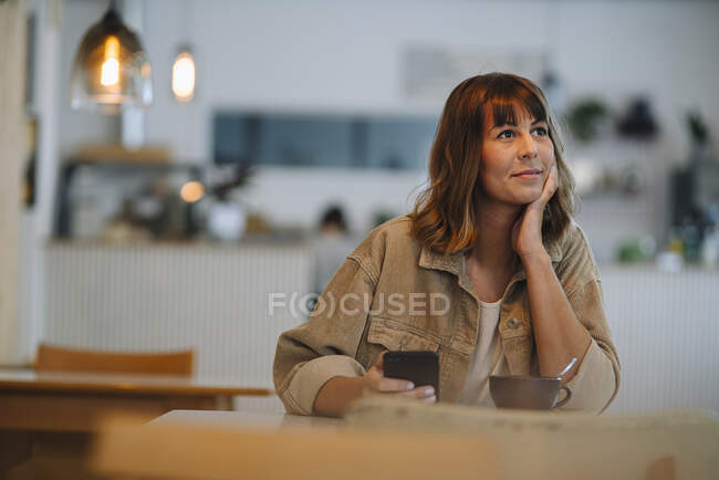 Propriétaire femelle avec la main sur le menton tenant le téléphone intelligent assis dans le café — Photo de stock
