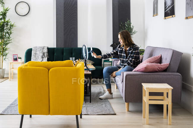 Mujer joven ajustando el teléfono móvil conectado con el flash del anillo mientras está sentado en el sofá en casa - foto de stock