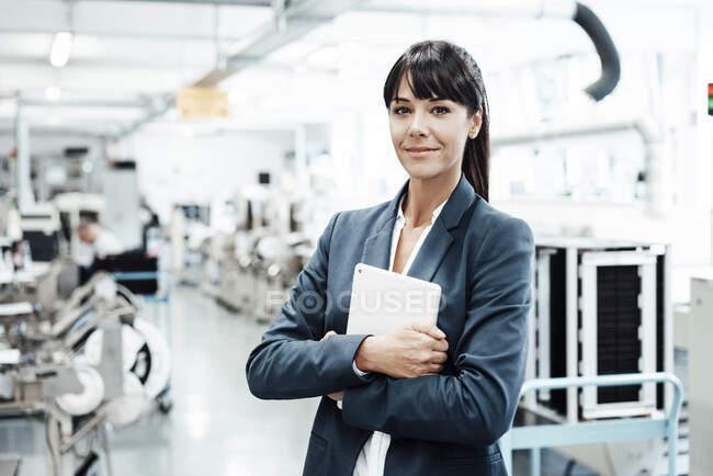 Mujer de negocios sonriente sosteniendo tableta digital mientras está de pie con los brazos cruzados en la industria - foto de stock