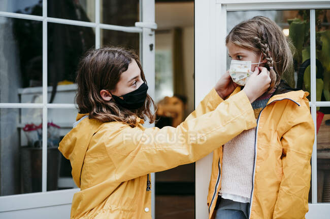 Sister adjusting face mask of sister while sitting against door — Stock Photo