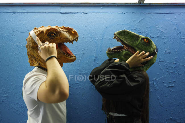 Amigos masculinos e femininos ouvindo música através de fones de ouvido enquanto usavam máscara de dinossauro contra a parede azul — Fotografia de Stock