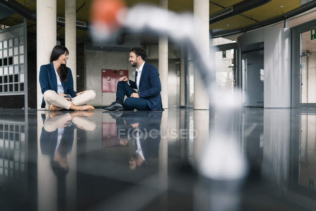 Улыбающиеся коллеги мужского и женского пола, обсуждающие, сидя на полу в офисном холле — стоковое фото