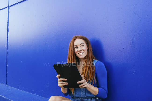 Усміхнена руда жінка використовує цифровий планшет, сидячи на синій стіні — стокове фото