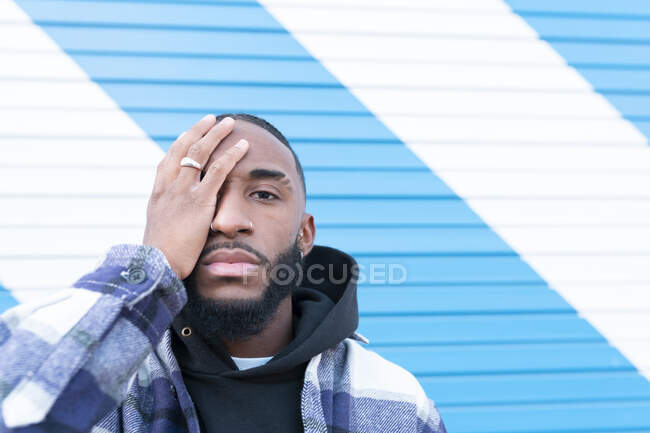 Male rapper covering eye with hand against striped blue and white corrugated wall — Stock Photo