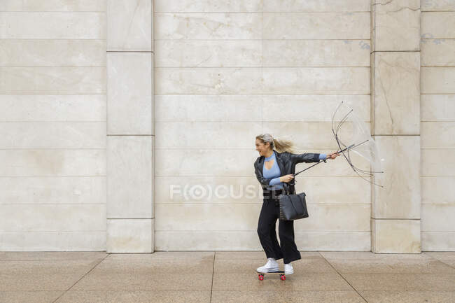 Cheerful female professional skateboarding while holding damaged umbrella against wall on rainy day — Foto stock