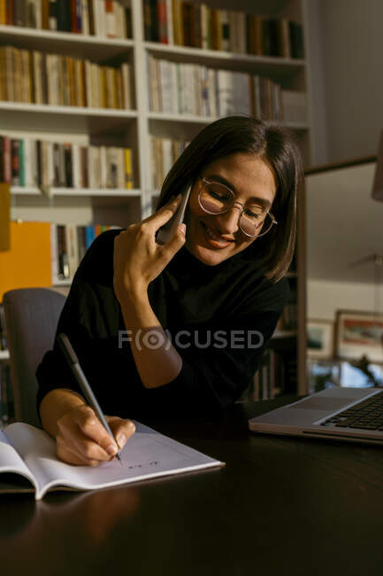 Усміхнена бізнес-жінка розмовляє на смартфоні, пишучи в книжках сидячи вдома. — стокове фото