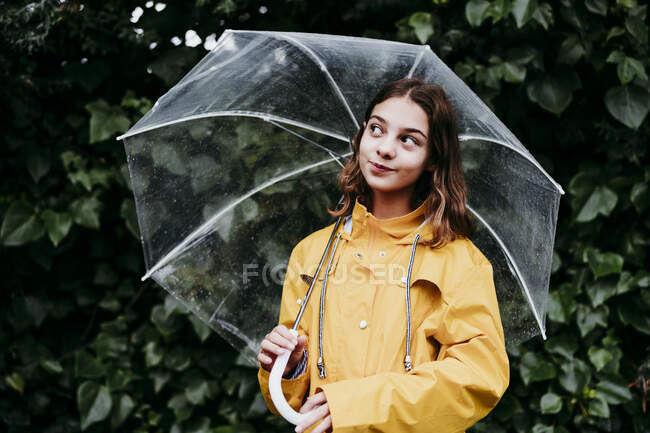 Lächelndes Mädchen im Regenmantel mit Regenschirm, das wegschaut, während es an der Laubwand steht — Stockfoto