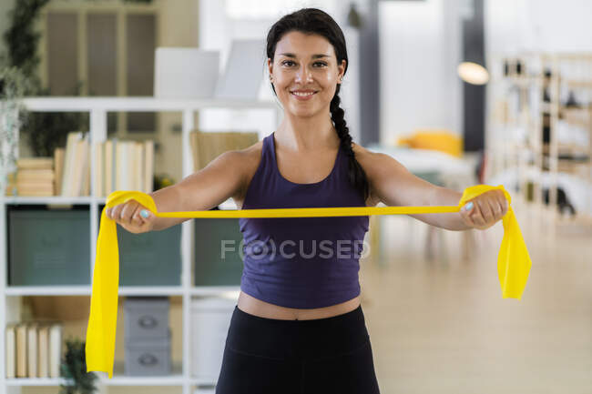 Giovane fitness femminile sorridente che si allunga con fascia di resistenza in piedi a casa — Foto stock