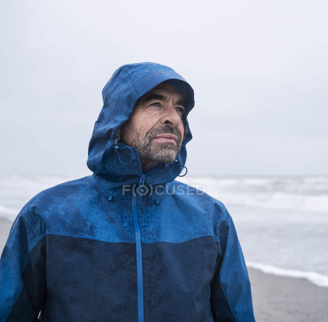 Pensativo homem maduro em capa de chuva azul em pé na praia contra o céu — Fotografia de Stock