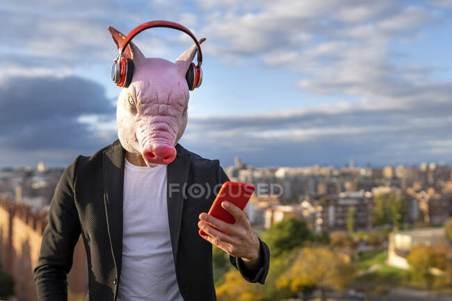 Geschäftsmann mit Schweinemaske und drahtlosen Kopfhörern mit Smartphone gegen den Himmel — Stockfoto