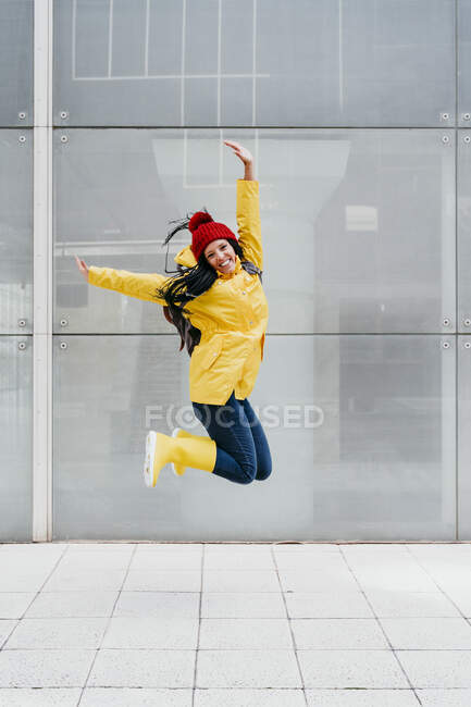 Happy woman jumping with hand raised on footpath against building exterior — Stock Photo