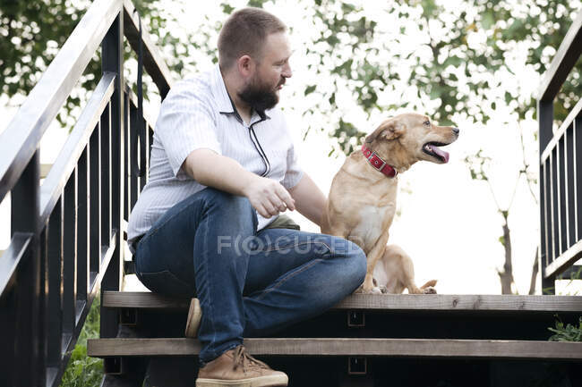Взрослый мужчина сидит с собакой на лестнице под открытым небом — стоковое фото