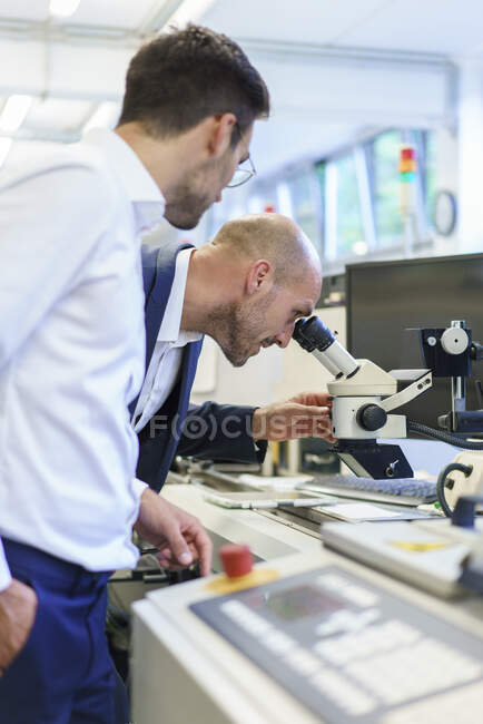 Joven técnico de pie por colega masculino mirando a través del microscopio en el laboratorio - foto de stock