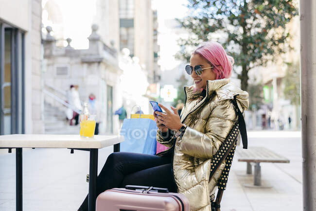 Sonriente mujer de pelo rosa usando el teléfono móvil mientras está sentado en el café de la acera - foto de stock
