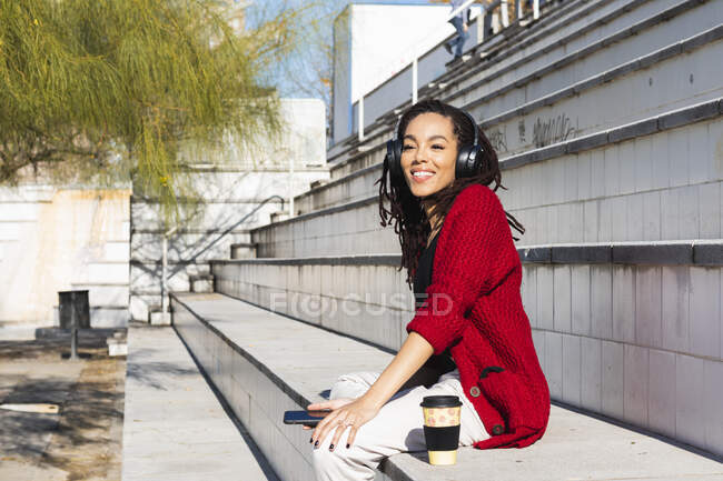 Smiling young female millennial sitting with smart phone and reusable cup at steps on sunny day — Stock Photo
