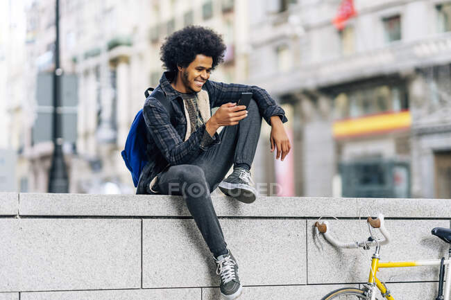 Sorrindo homem com cabelo encaracolado usando telefone celular enquanto sentado na parede de retenção na cidade — Fotografia de Stock