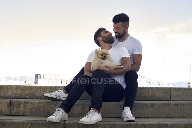 Улыбающаяся гей-пара с собакой, сидящей на ступеньках над небом — стоковое фото