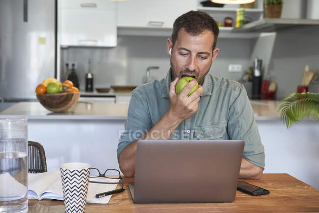 Guapo freelancer masculino comiendo manzana fresca mientras trabaja en casa con computadora portátil - foto de stock