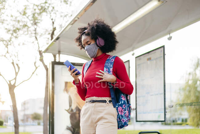 Jeune femme portant un casque et un masque protecteur en utilisant un téléphone intelligent tout en restant debout à l'arrêt de bus — Photo de stock