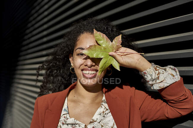 Young woman covering face with autumn colored leaf against shutter on sunny day — Stock Photo