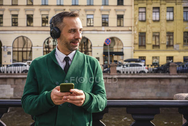 Empresario con auriculares mirando hacia otro lado mientras usa el teléfono móvil en la ciudad - foto de stock