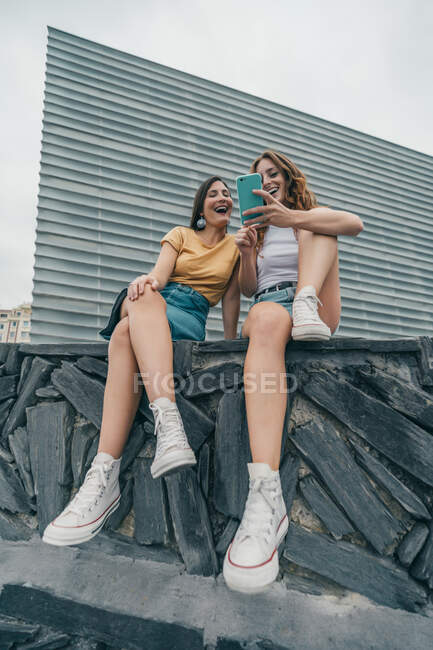 Sonrientes amigas usando el teléfono móvil mientras están sentadas contra la pared - foto de stock