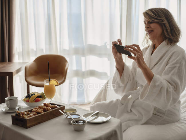 Улыбающаяся блондинка пожилая женщина фотографирует завтрак через смартфон, сидя на кровати в номере отеля — стоковое фото