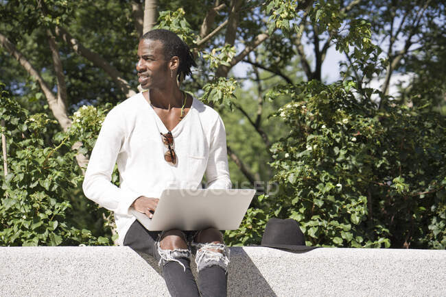 Mann benutzt Laptop während er auf Stützmauer im Park sitzt — Stockfoto