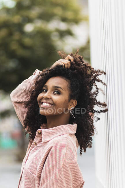 Afro mujer sonriendo con la mano en el pelo mientras está de pie en la ciudad - foto de stock