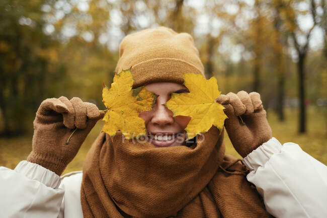 Happy young woman wrapped in scarf covering eyes with autumn leaf in public park — Stock Photo