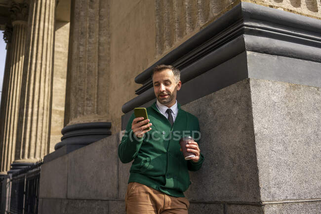Businessman with coffee cup using mobile phone while leaning on pillar of Kazan Cathedral at Saint Petersburg, Russia — Stock Photo